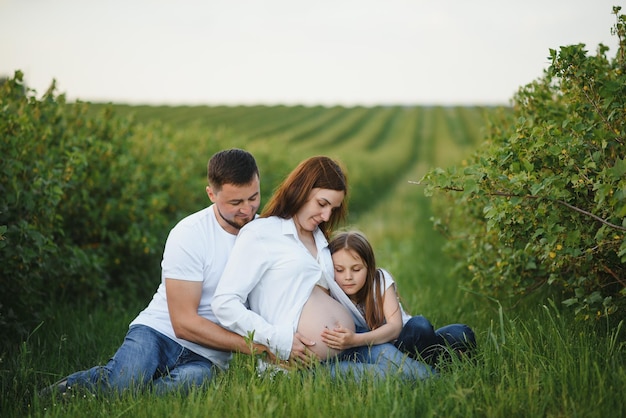 Beautiful pregnant brunette mom and dad and dauther in white clothes look at each other in nature