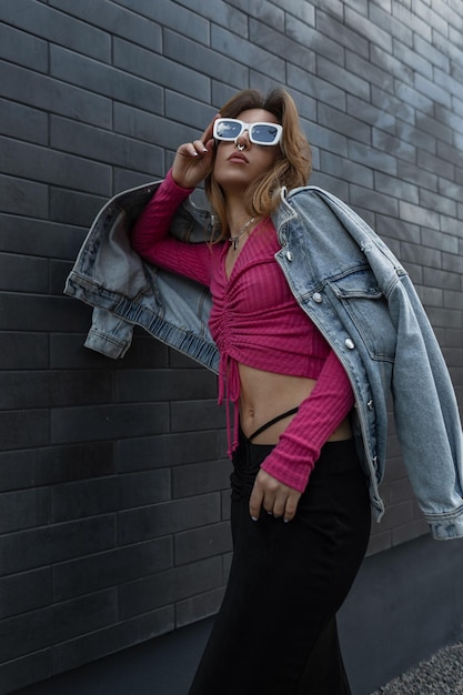 Beautiful redhaired fashion model girl with a nose piercing with sunglasses in fashionable denim clothes with jacket pink top and stylish black skirt stands and poses near a black brick wall