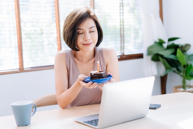 Photo beautiful young asian woman celebrating birthday at home during pandemic quarantine timeher blows cake with friends on through video call virtual party with laptop computercoronavirus outbreak 2020