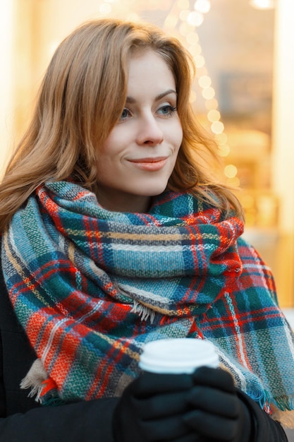 Beautiful young girl model in fashion clothes with a vintage plaid scarf walks on the street and drinks coffee