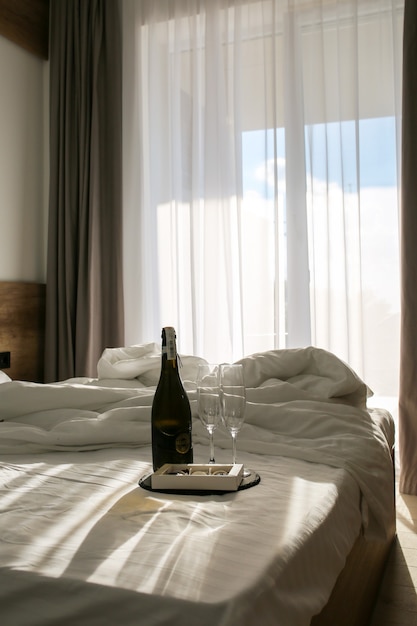 Bedroom with a spread bed White linens on the bed with champagne and glasses Summer
The suns rays fall on the bed Morning in the bedroom with champagne and sweets Bed with champagne and glasses