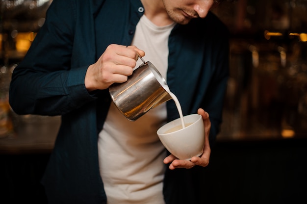 Brunet bartender pouring a milk into the white ceramic cup of coffee