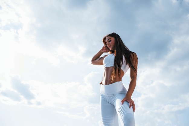 Brunette fitness woman stands outdoors in white sportive clothes against cloudy sky at daytime