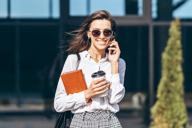 Business woman walking and talking on the cellphone outdoors near modern business center with notebook