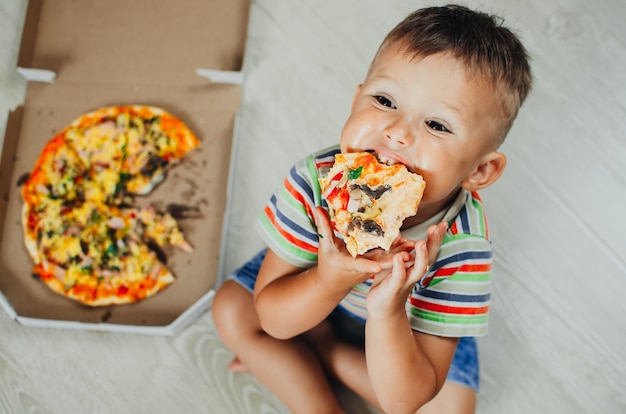 Charming boy sitting on the floor eating pizza top view