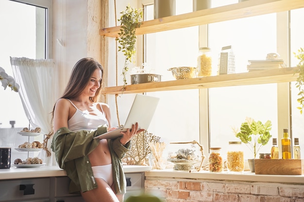 Charming girl. Pleased brunette leaning on her furniture while working at her laptop in the kitchen