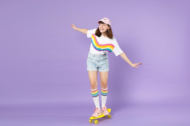 Photo cheerful teen girl in vivid clothes riding yellow skateboard spreading hands looking aside isolated on violet wall background in studio. people sincere emotions lifestyle concept. mock up copy space.