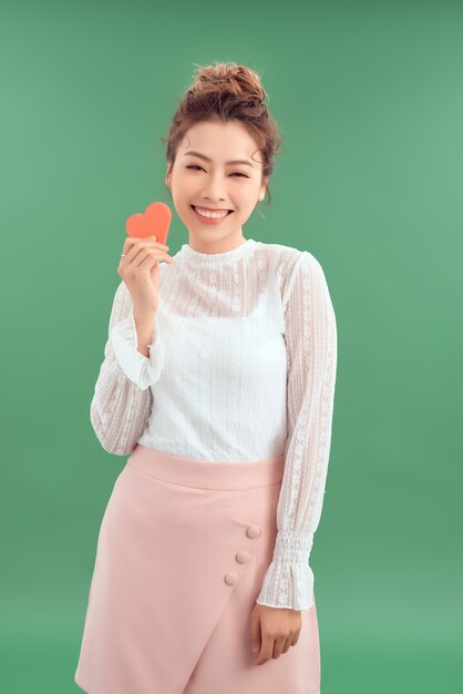 Photo cheerful young asian woman holding red heart paper isolated over green background.