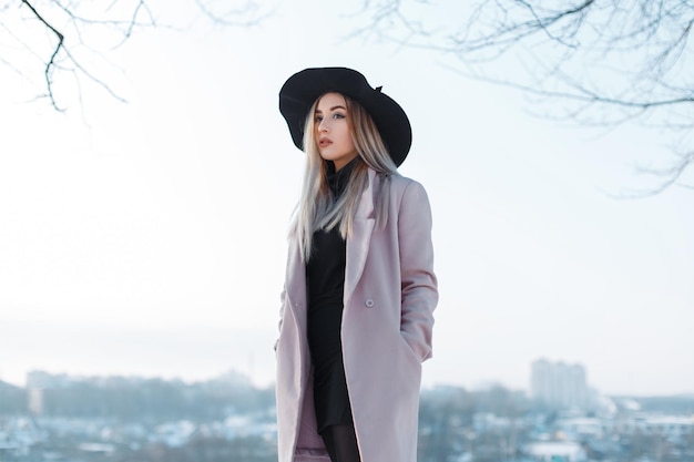 Chic beautiful fashion elegant woman with black hat and trendy stylish coat stands outdoors in winter snowy day