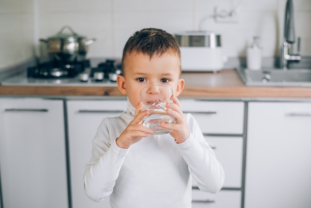 a child drinks clean water from a glass on the background of white kitchen furniture