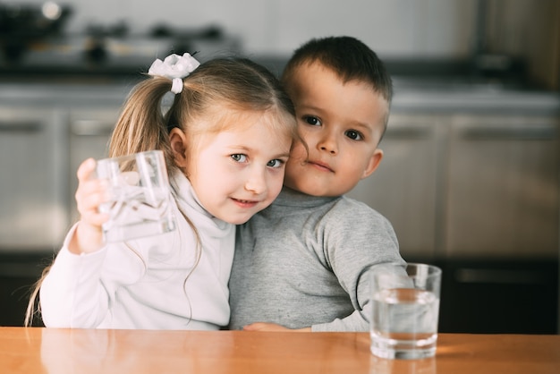 Children boy and girl in the kitchen drinking water from glasses, hugging and smiling very sweet