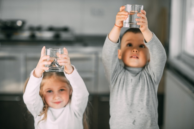 Children boy and girl in the kitchen holding glasses raised above his head very sweet