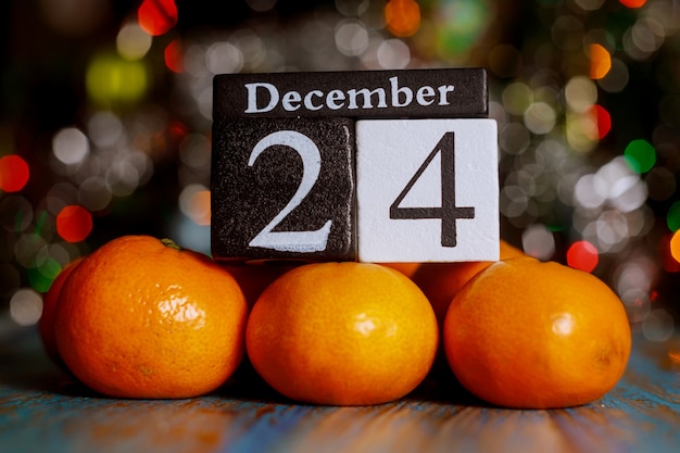 Photo christmas eve, december 24 th wooden cube calendar with tangerines on christmas tree lights background.