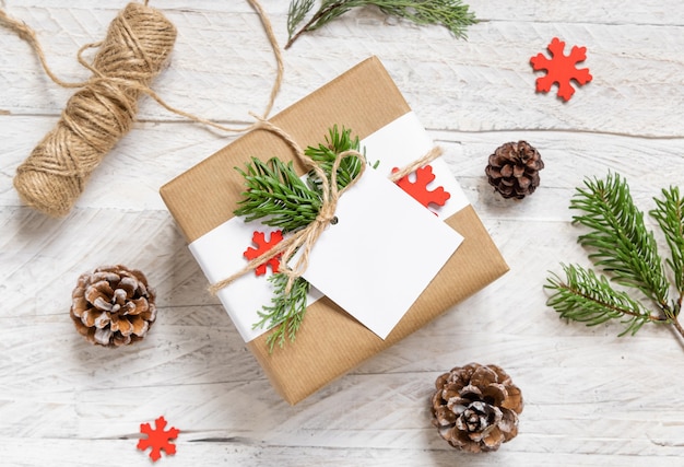 Christmas wrapped present with square paper gift tag on a white table with fir tree branches and decorations top view. Rustic winter composition with blank Gift tag Mockup, copy space