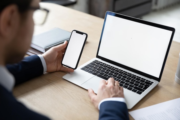 Photo close up back view of male employer sit at desk using modern laptop and smartphone gadget businessman consult client work on computer and cellphone device at workplace white mockup screen