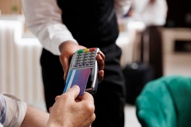 Close-up of elderly man using credit card to make financial transaction in cosy lounge area. Detailed shot showing cashless payment done at hotel lobby with client and african american waiter.