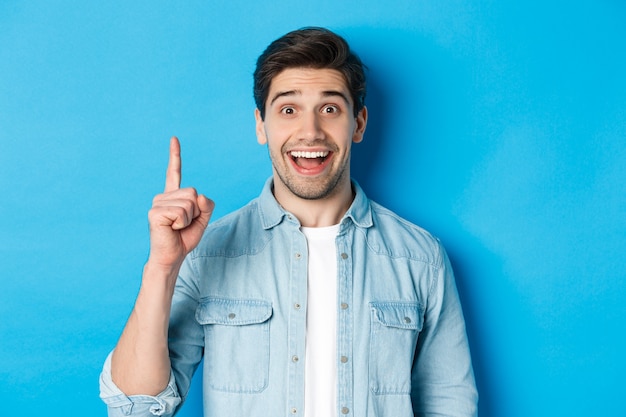 Close-up of handsome bearded guy smiling, showing finger number one, standing over blue wall