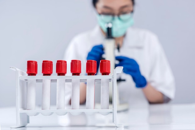 Photo closeup view of the medical tube rack with asian researcher woman with face mask and glasses using a microscope on the lab