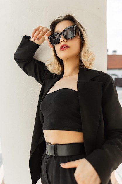 Cool beautiful stylish model woman model with cool fashion black sunglasses in fashion elegant business clothes with black blazer and top stands and poses near a vintage white column