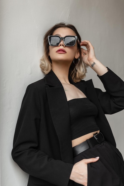 Cool elegant fashion young woman with stylish eyewear in black fashionable business outfit with top and blazer poses near a white wall on the street