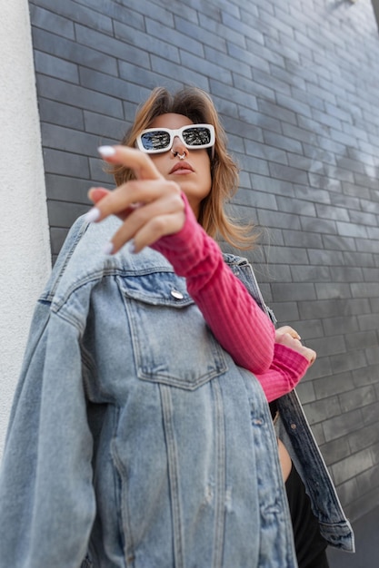 Cool stylish young woman hipster model with white cool sunglasses in fashion urban outfit with jeans jacket and pink sweatshirt poses near a brick black wall in the city