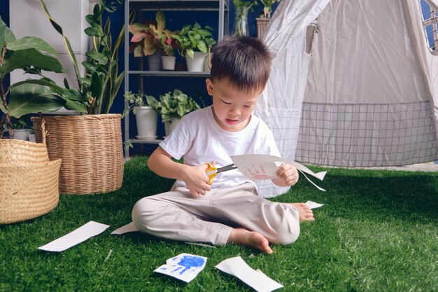 Photo cute asian kindergarten boy cutting a piece of paper, introduce scissor skills for toddler