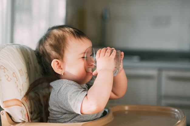 Cute little girl sitting in a baby chair in the kitchen and drinking water