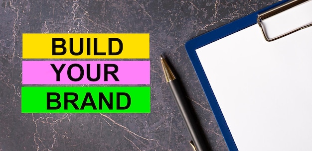 On a dark marble background a tablet with paper pen and three bright multicolored strips of paper with the text BUILD YOUR BRAND