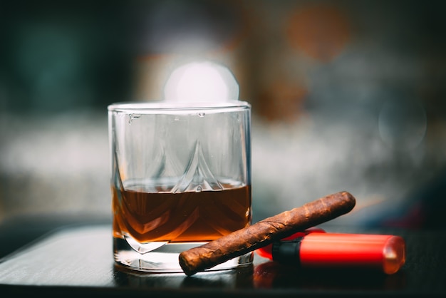 Decanter of whiskey and a glass with cuban cigar and golden lighter on a wooden table. Angle view alcohol and tobacco