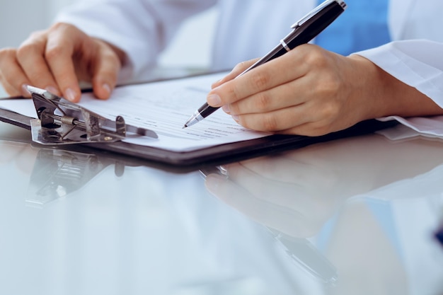 Doctor woman filling up medical form while sitting at the table, close-up of hands.