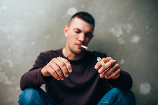 A drug addict with a syringe in his hand and a cigarette in his teeth is sitting on the floor after ...