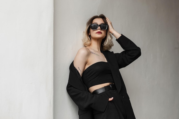 Elegante modieuze zakelijke glamour vrouw model met fashion zonnebril in zwarte kleding met top en stijlvolle zwarte blazer staat in de buurt van een muur op straat