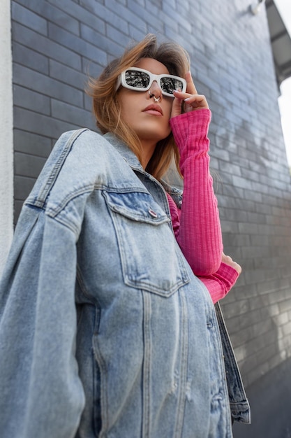 Fashion redhead beauty woman model with blue fashionable denim jacket in pink sweatshirt stands and puts on a white trendy sunglasses on the street Urban female outfit look