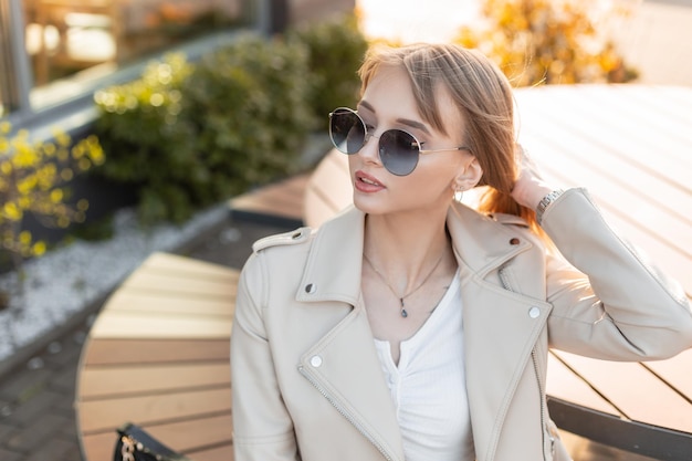 Fashion street portrait of beauty hipster woman with fashionable cool round sunglasses in leather rock stylish jacket sits and rest on wooden bench on the street at sunset