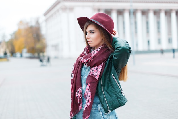 fashionable beautiful young girl in a stylish vintage burgundy hat with a scarf in a fashion green leather jacket walks in the city