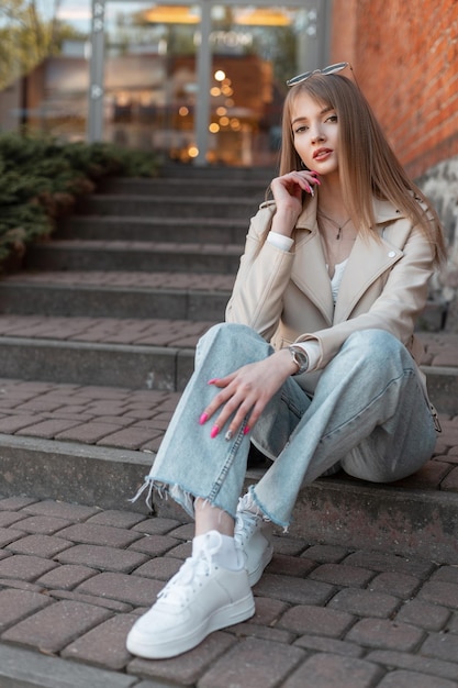 Fashionable young beautiful woman in trendy autumn clothes with fashion white leather jacket and blue vintage jeans with sneakers sits on the steps near a mall in the city