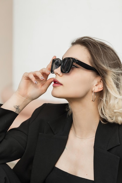 Fashionable young business elegant woman model with trendy modern sunglasses in black outfit with top and blazer sits on the street Urban female look clothes