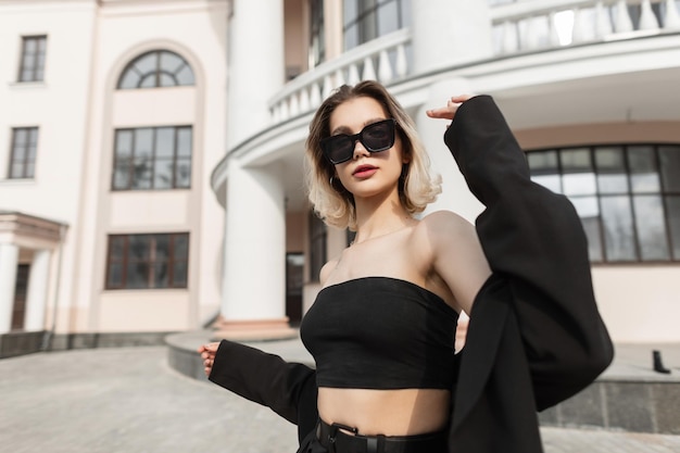 Fashionable young business woman model with stylish modern sunglasses in black fashion elegant clothes with top and blazer walks on the street near a vintage building Urban style and fashion