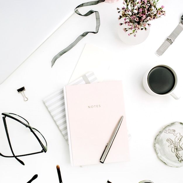 Feminine modern home office desk with pastel pink notebook, glasses, coffee cup, wildflowers. Flat lay, top view