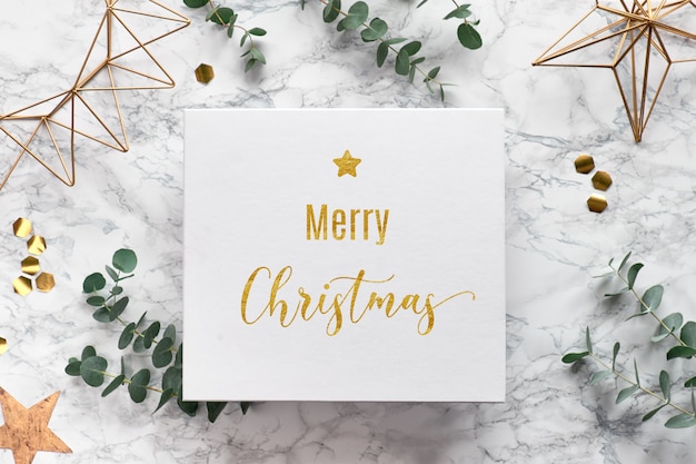 Photo flat lay on white marble background with text "merry christmas" on canvas. christmas frame with fresh eucalyptus twigs and golden geometric decorations - hexagons, trinkets and wire shapes.