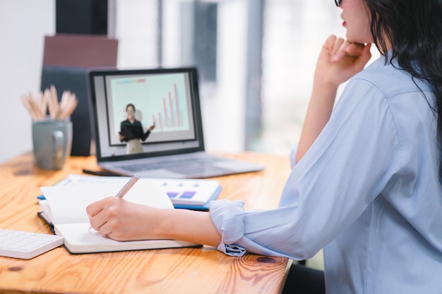 Photo focused asian businesswoman wearing headphones is taking notes in a notebook while watching a webinar video course serious female student listening to the lecture to study online through elearning