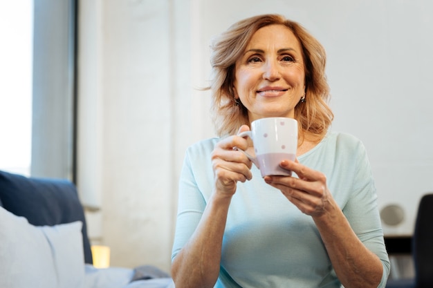 Freshly-made coffee. Smiling short-haired mature lady being happy with her peaceful morning while drinking coffee and building plans