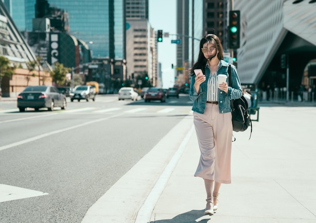 Photo full length cheerful young asian woman using mobile phone in sunny city street and drinking take away coffee in paper cup. female employee with backpack walking on road in busy business area traffic