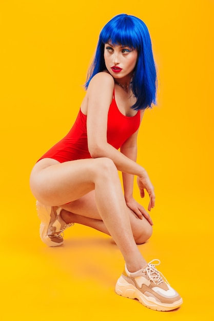 Full length portrait of an attractive confident young funky girl with blue hair wearing swimsuit sitting isolated over yellow wall