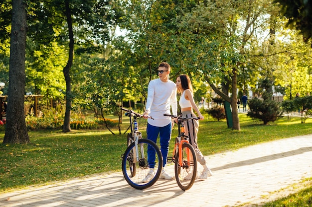 Full length portrait of a beautiful young man and woman standing in the park with bicycles