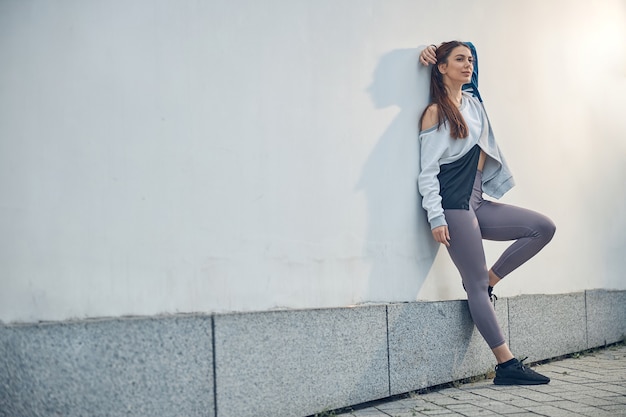 Full-length portrait of a dreamy slender young woman leaning her back against a wall outdoors