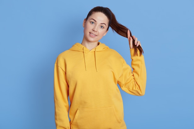 Foto glimlachende jonge vrouw in casual gele hoodie poseren geïsoleerd op blauwe muur, met paardenstaart, camera kijken met charmante glimlach, positieve emoties uitdrukken.