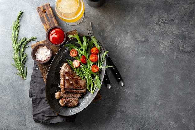 Grilled beef steak well done with fresh vegetable salad and a glass of light beer on a black plate on the dark gray background top view copy space for text