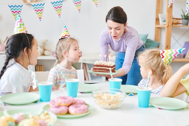 Group of cute little girls and pretty young woman blowing candles on birthday cake during home celebration by festive table