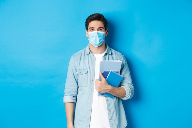 Guy student in medical mask looking happy, holding notebooks, standing over blue wall